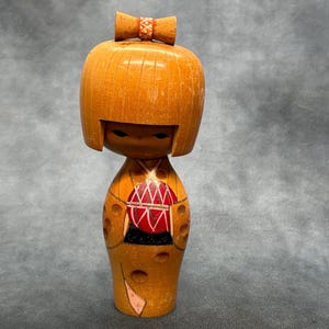 May include: A wooden Kokeshi doll with a light brown finish. The doll has a rounded head with a small bow and a painted face. The body features a red and black design. This traditional Japanese doll is a collectible item.