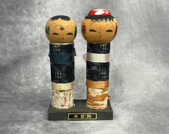 May include: Two traditional Japanese wooden dolls, known as kokeshi, with painted faces and colorful clothing. The dolls are standing on a black wooden base with the text "木曾路" in gold lettering.