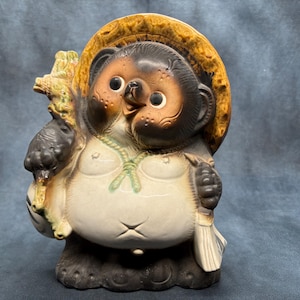 Peut inclure: Figurine de Tanuki avec un chapeau de paille, tenant une bouteille de saké et un billet à ordre. La statue en céramique a un gros ventre, une expression amicale et est peinte dans des tons de marron, beige et blanc. Un objet d'art populaire japonais traditionnel.