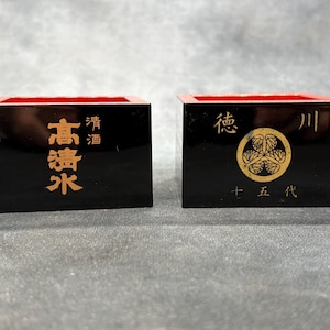 May include: Two black wooden sake cups with red interiors. One cup has gold Japanese characters that read "Takashimizu" and "Seishu". The other cup has gold Japanese characters and a gold emblem.