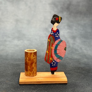 May include: A wooden Japanese geisha figurine with a cylindrical holder. The geisha is dressed in a blue kimono with red and gold accents, holding a pink fan. The figurine and holder are mounted on a rectangular wooden base.