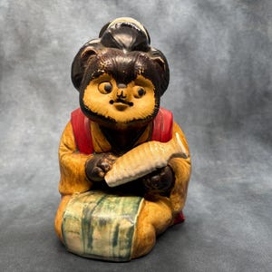 Peut inclure: Statue en céramique de Tanuki, une figurine traditionnelle japonaise de chien viverrin. La statue est assise, portant un gilet rouge et tenant un poisson. La figurine a un visage détaillé et est peinte dans des tons de marron, jaune et vert.