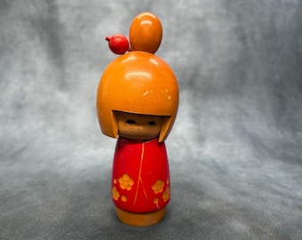 May include: Wooden Kokeshi doll with a red dress and orange floral accents. The doll has a round head with a small red bird on top. The doll's face is simple, with small eyes and a painted mouth.