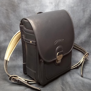 May include: A black leather backpack with a large flap and a buckle closure. The backpack has a smaller pocket on the front and a cream-colored strap.