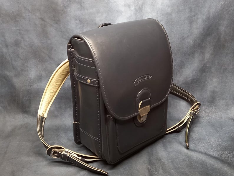 May include: A black leather backpack with a large flap and a buckle closure. The backpack has a smaller pocket on the front and a cream-colored strap.