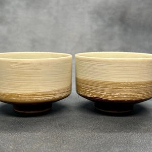 May include: Two ceramic tea bowls featuring a horizontal brushed texture. The upper portion is a creamy off-white, transitioning into a warm tan and dark brown base. The bowls have a wide, open rim and a small, raised circular foot at the bottom.