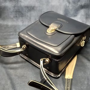 May include: A black leather camera bag with a gold-toned clasp and a long adjustable strap. The bag has a flap closure and a small pocket on the front.