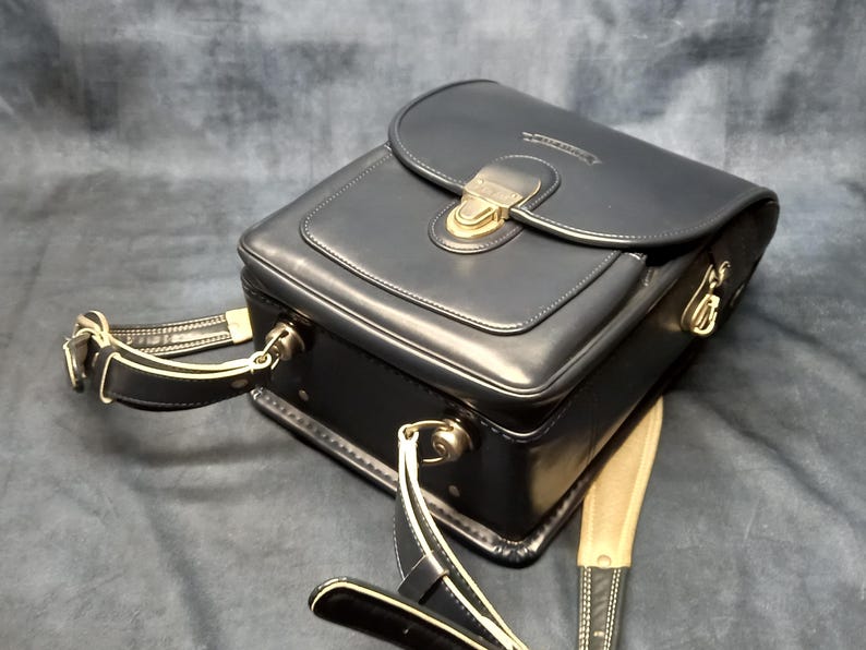 May include: A black leather camera bag with a gold-toned clasp and a long adjustable strap. The bag has a flap closure and a small pocket on the front.