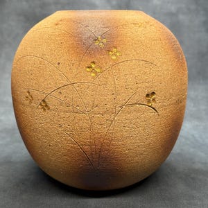 May include: A round, brown ceramic vase with a textured surface. The vase features etched floral and leaf designs, with small, yellow-gold flower accents. The top rim is slightly darker in colour.
