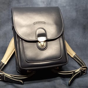 May include: Black leather backpack with a gold buckle and a white strap. The backpack has a small pocket on the front with the text "RED  CRAFT" embossed on it.