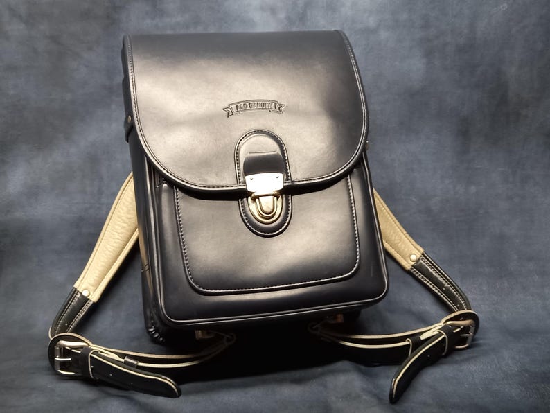 May include: Black leather backpack with a gold buckle and a white strap. The backpack has a small pocket on the front with the text "RED  CRAFT" embossed on it.