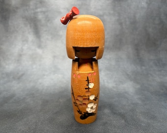 Puede incluir: Una muñeca Kokeshi de madera con cabeza redondeada y cuerpo cilíndrico. La muñeca está pintada con un acabado marrón claro y presenta un lazo rojo en la parte superior. El cuerpo está decorado con diseños florales y de ramas en tonos rosa, blanco y marrón.