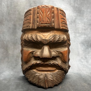 May include: A hand-carved wooden sculpture depicting a bearded face with a prominent brow and a textured hat. The piece features deep, detailed chisel marks across the hair, beard, and headwear, showcasing natural wood grain and varied brown tones.