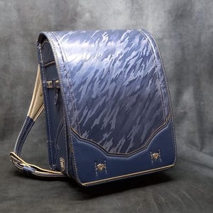 May include: A navy blue and silver patterned school backpack with a structured design. The backpack features a flap closure, a top handle, and adjustable shoulder straps. The bottom section is a darker navy blue with decorative accents.