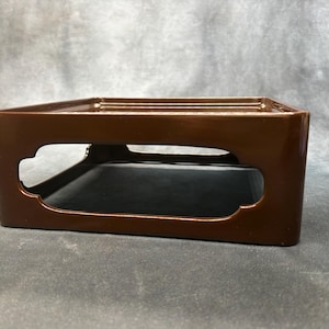 May include: A square, brown, glossy lacquerware stand with a flat top surface and a hollowed-out centre featuring decorative, rounded cutouts on each side. The base is open, providing a stable platform for display with a smooth, reflective finish.