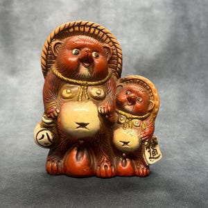 Peut inclure: Une figurine en céramique représentant deux tanukis au pelage brun-roux, au ventre crème et portant des chapeaux de paille. Le plus grand tient une bourse ronde marquée du caractère japonais 八, le plus petit tient une étiquette avec le caractère 道.