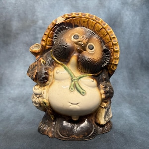 Peut inclure: Une figurine en céramique Tanuki avec une finition émaillée brune et crème. Le personnage porte un grand chapeau couleur paille, transporte une bouteille de saké et un registre, et possède un cordon vert noué autour de la poitrine.