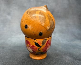 May include: A wooden decorative figurine with a rounded top and a cup-shaped base. The top is light brown with darker brown swirls and black eyes. The base is orange with red and white floral designs.