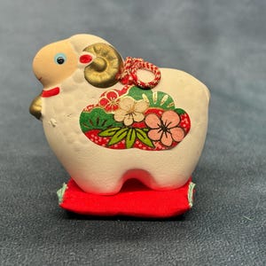 Puede incluir: Una figura de carnero de cerámica color crema con un cuerno pintado de dorado, un cordón trenzado rojo y blanco, y un patrón floral decorativo en el costado en tonos rojo, verde y rosa. El carnero se asienta sobre un pequeño cojín de tela rojo.