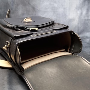 May include: A black leather satchel bag with a flap closure and a gold buckle. The bag is open and the inside is lined with a beige fabric.
