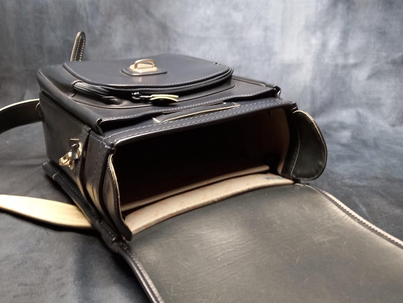 May include: A black leather satchel bag with a flap closure and a gold buckle. The bag is open and the inside is lined with a beige fabric.