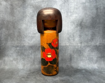 Puede incluir: Muñeca Kokeshi japonesa de madera. La muñeca presenta una cabeza marrón oscuro y un cuerpo cilíndrico decorado con diseños florales rojos y naranjas. La muñeca mide unos 15 cm de altura.