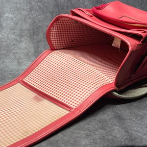 May include: A red leather school backpack with a checkered lining. The backpack is open and the lining is visible. The backpack has a strap and a handle.