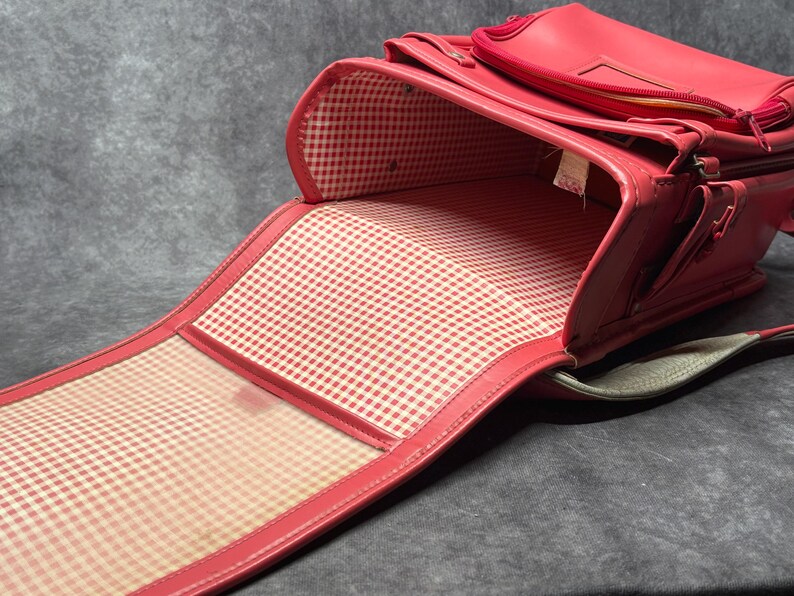 May include: A red leather school backpack with a checkered lining. The backpack is open and the lining is visible. The backpack has a strap and a handle.