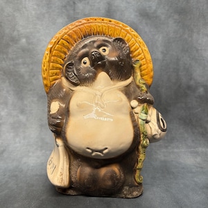 May include: A ceramic tanuki statue, a traditional Japanese raccoon dog figure. The statue has a large belly, a straw hat, and holds a sake bottle and a bag. The colors are brown, tan, and cream.