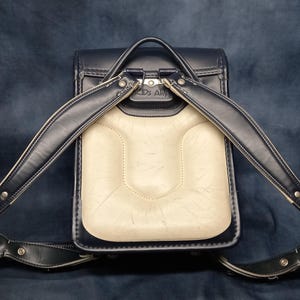 May include: A black leather backpack with a white leather front panel and silver hardware. The backpack has two adjustable straps and a top handle.