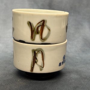 May include: Two stacked, off-white ceramic cups with brown and green Japanese characters. The cups have a cylindrical shape with a black base and a blue logo on the side. The cups are likely for tea or sake.
