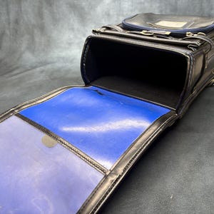 May include: Black leather school bag with a rectangular shape, open to reveal a blue interior. The bag has a top flap, a handle, and silver-colored hardware. The interior is a vibrant blue color.
