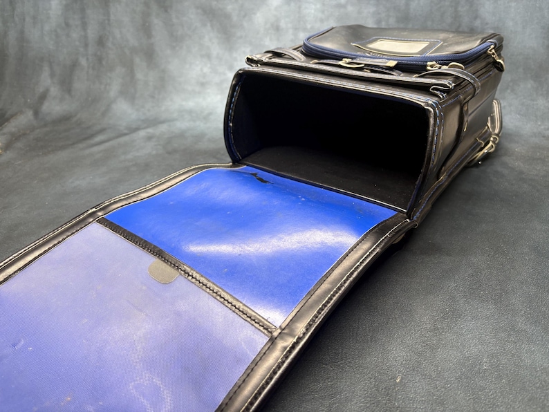 May include: Black leather school bag with a rectangular shape, open to reveal a blue interior. The bag has a top flap, a handle, and silver-colored hardware. The interior is a vibrant blue color.