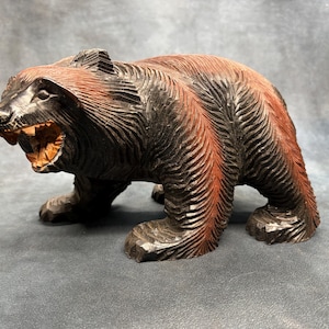 Puede incluir: Una figura de oso de madera tallada en pose de rugido. El oso es principalmente negro con acentos marrón rojizo a lo largo de su espalda. La boca está abierta, revelando dientes tallados y un interior de madera más clara.