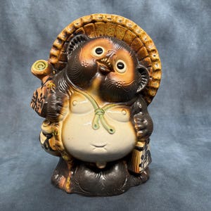 Peut inclure: Une statue en céramique de Tanuki, une figure traditionnelle japonaise de chien viverrin. La statue présente un grand ventre, un chapeau de paille et tient une bouteille de saké. Les couleurs sont marron, beige et crème, avec des traits de visage détaillés et un sourire.