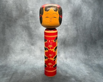 May include: A wooden Japanese Kokeshi doll with a smiling face and black hair. The doll's body is red with a floral design in red and black. The head is orange and black.