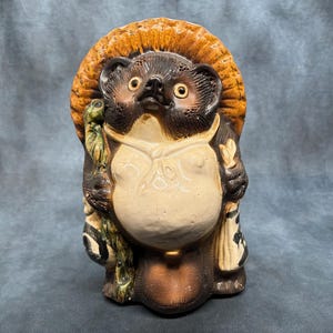 Peut inclure: Statue en céramique de Tanuki, une figure traditionnelle japonaise de chien viverrin. La statue présente un schéma de couleurs beige, marron et noir, avec un grand ventre, tenant une bouteille de saké et un sac. Elle porte un chapeau de paille et a une expression amicale.