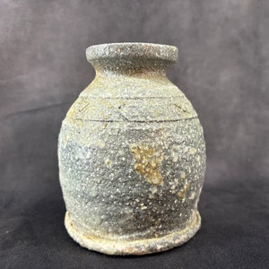 Puede incluir: Un pequeño jarrón de cerámica gris con un esmalte moteado y una superficie rugosa y texturizada. El jarrón tiene un cuerpo redondeado y una abertura ligeramente acampanada.