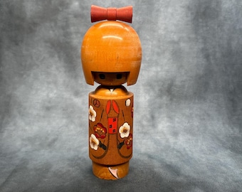 Puede incluir: Muñeca Kokeshi de madera con lazo rojo, cabeza naranja y cuerpo cilíndrico. El cuerpo está decorado con motivos florales y geométricos en rojo, blanco y marrón. Artesanía popular japonesa tradicional.