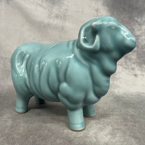 Puede incluir: Una figurita de cerámica de oveja azul claro con un vellón rizado y una cabeza prominente con cuernos.
