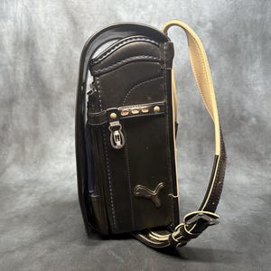 May include: Black leather Puma school backpack with a beige shoulder strap. The backpack has a rectangular shape with a flap closure, a front pocket, and a Puma logo. The bag has silver hardware and stitching.