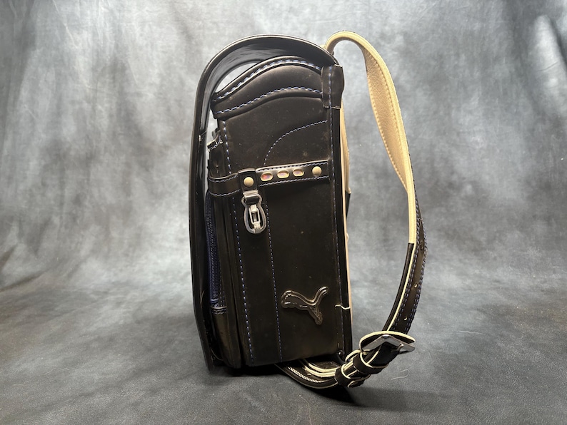 May include: Black leather Puma school backpack with a beige shoulder strap. The backpack has a rectangular shape with a flap closure, a front pocket, and a Puma logo. The bag has silver hardware and stitching.