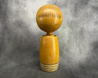 May include: A wooden doll with a round head and a cylindrical body. The head is painted brown with a pattern of brown lines. The body is painted brown with a band of white stripes around the bottom.
