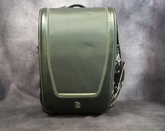 May include: A dark green backpack with a textured fabric exterior and a single zippered pocket on the front. The backpack has a black strap and a silver buckle.