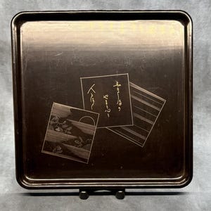 May include: A square, dark brown tray with rounded corners. The tray features three rectangular designs with gold-colored illustrations and Japanese text. The tray is displayed on a black stand.