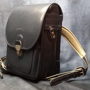 May include: A black leather backpack with a flap closure and a buckle. The backpack has a brown leather strap and a small pocket on the front.