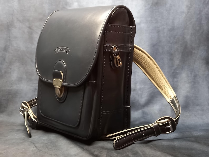 May include: A black leather backpack with a flap closure and a buckle. The backpack has a brown leather strap and a small pocket on the front.