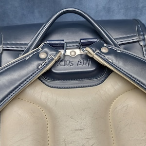 May include: A navy blue leather backpack with a beige leather interior. The backpack has two adjustable straps and a metal buckle. The backpack has the text "KIDS AMI" printed on the front.