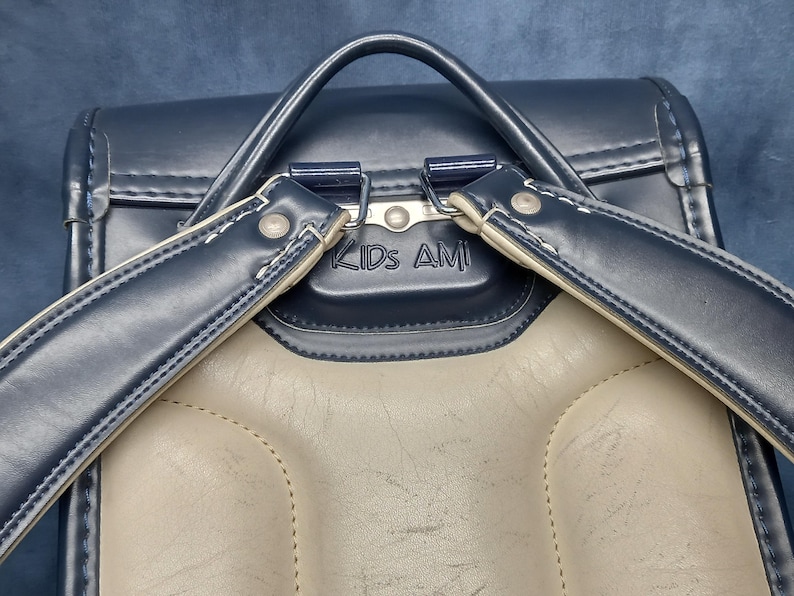 May include: A navy blue leather backpack with a beige leather interior. The backpack has two adjustable straps and a metal buckle. The backpack has the text "KIDS AMI" printed on the front.