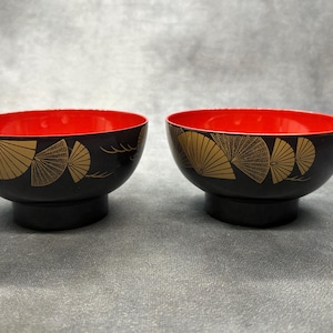 May include: Two black lacquer bowls with a red interior. The exterior features a gold fan design. The bowls are round with a pedestal base. The bowls are likely for serving food.